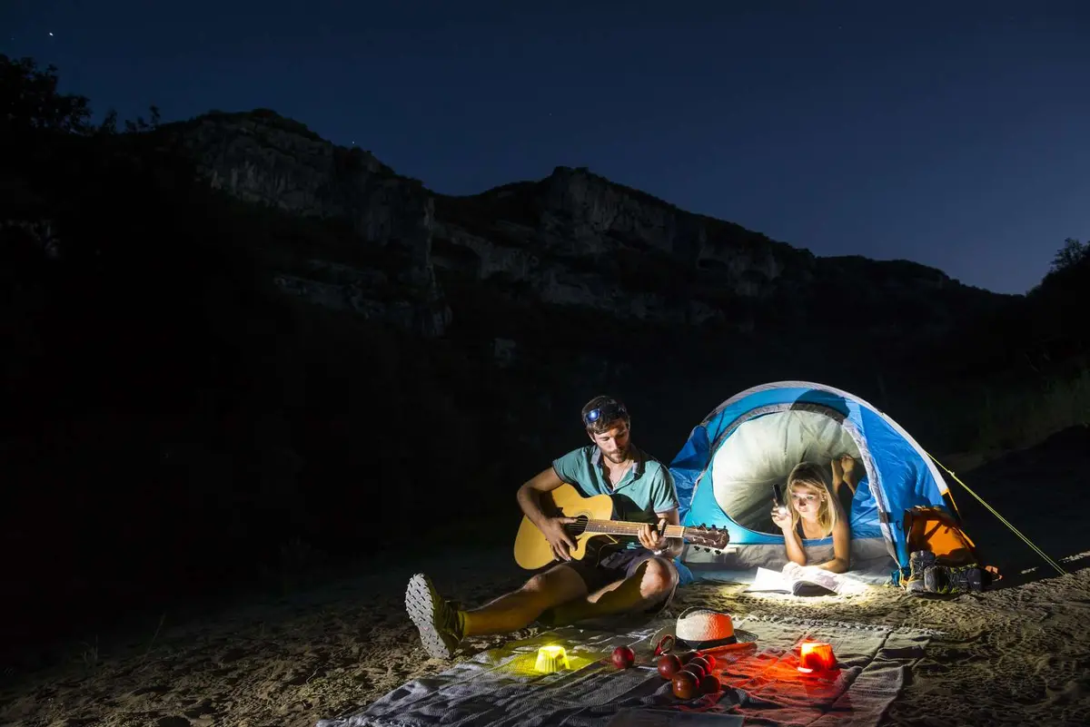 Une tente éclairée sous les falaises à la tombée de la nuit, un campeur joue de la guitare