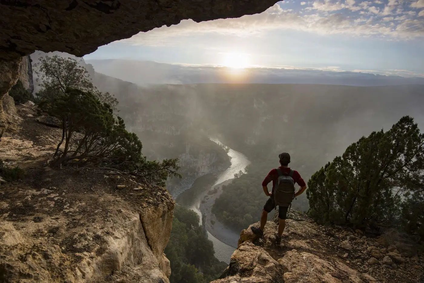 Depuis l'entrée d'une grotte, un randonneur contemple les méandres de l'Ardèche au coucher du soleil
